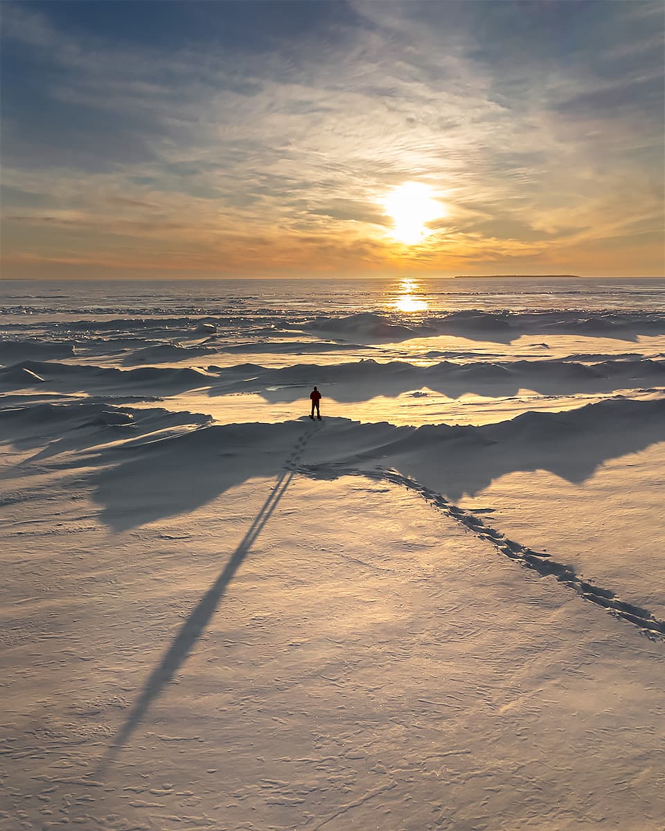 Long shadow of man standing on frozen mounds of lake ice during sunset.